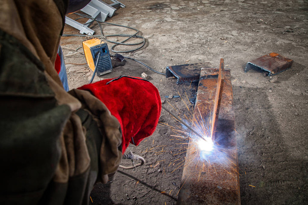 Nahaufnahme eines Bauarbeiters in brauner Arbeitskleidung schweißt in der Fabrik ein Metallprodukt mit Lichtbogenschweißmaschine, vielen Werkzeugen und Beschränkungen, hellen Lichtern und Funken - Foto, Bild