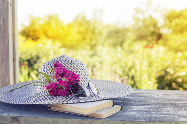Female hat with glasses, books and pink flowers on blurred nature background - Фото, зображення