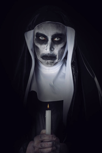 lähikuva pelottavasta pahasta nunnasta, jolla on tyypillinen mustavalkoinen tapa, mustaa taustaa vasten, kynttilä käsissään
 - Valokuva, kuva
