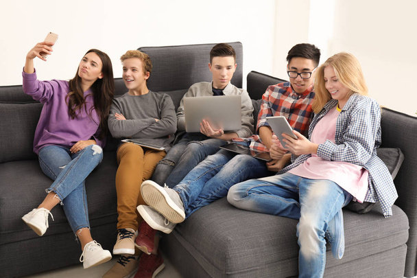 Group of teenagers with modern devices taking selfie while sitting on sofa indoors - Фото, зображення