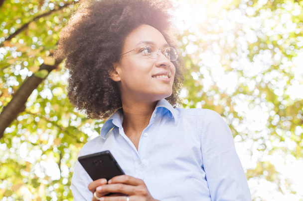 Εύθυμη νεαρή μαύρη γυναίκα που χρησιμοποιούν Smartphone στη φύση. Πορτραίτου, φοράει γυαλιά, βλέπουν την πλευρά, φως του ήλιου φωτοβολίδα στο παρασκήνιο - Φωτογραφία, εικόνα