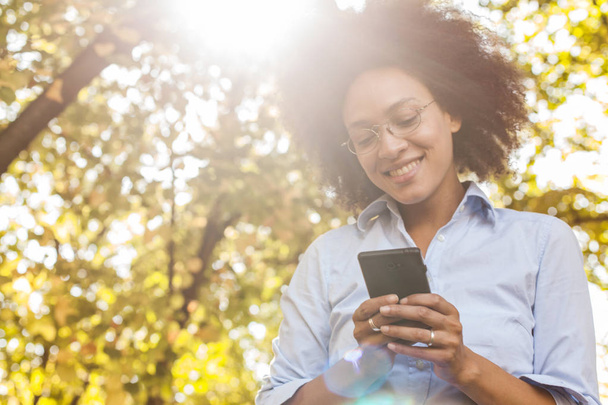 Hermosa mujer negra joven usando el teléfono en la naturaleza. Retrato al aire libre, lleva gafas, mira el teléfono, destello de luz solar en el fondo
 - Foto, Imagen