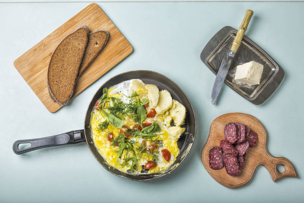 El desayuno de la tortilla con hortalizas, los tomates cherry y la albahaca en la sartén y los bocadillos con la mantequilla, el salami, el pan grueso
 - Foto, Imagen