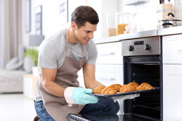 Man taking baking tray with delicious croissants out of electric oven in kitchen - Φωτογραφία, εικόνα