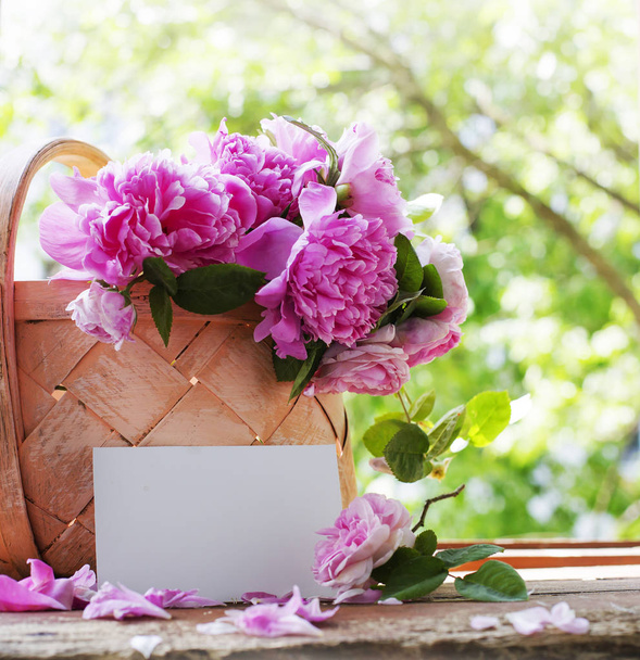 bouquet of fresh cut pink peonies in basket on garden table with empty card - Fotó, kép
