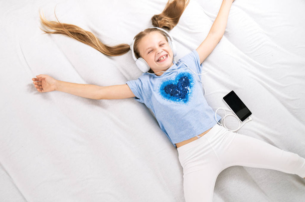 Music lover: The girl stretches out on a big white bed and listens to music in big white headphones. - Fotó, kép
