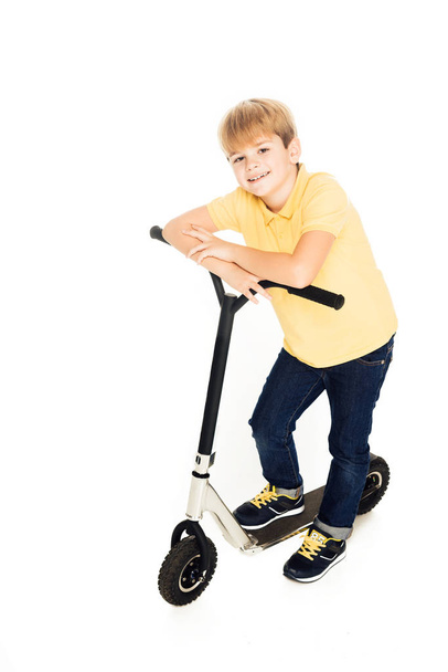 bonito menino feliz inclinando-se para scooter e sorrindo para a câmera isolada no branco
 - Foto, Imagem