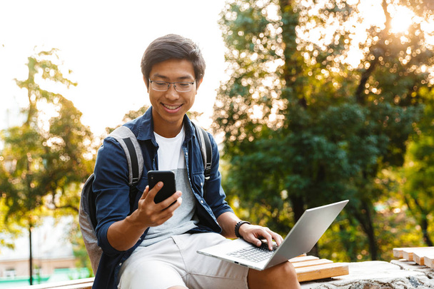 Smiling asian male student in eyeglasses using smartphone while sitting on bench in park with laptop computer - Фото, изображение