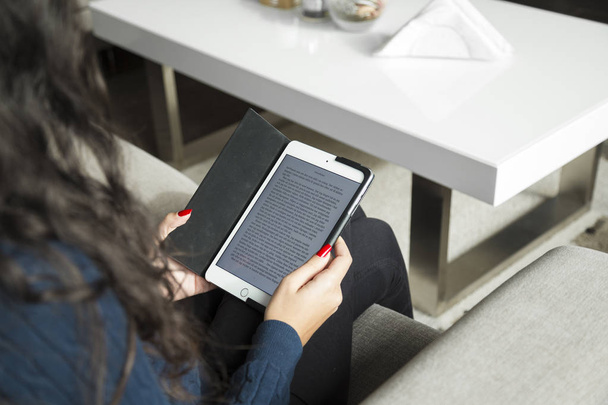 A young woman sitting in the living room on a couch and reading a book on her iPad. - Foto, afbeelding