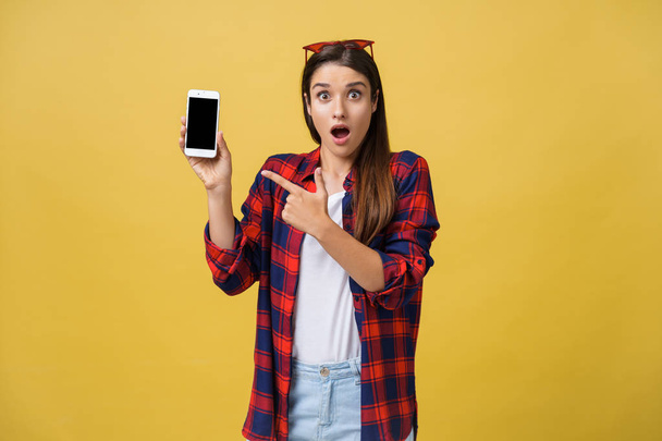 Photo of emotional shocked young pretty woman standing isolated over yellow background using mobile phone looking aside. - 写真・画像