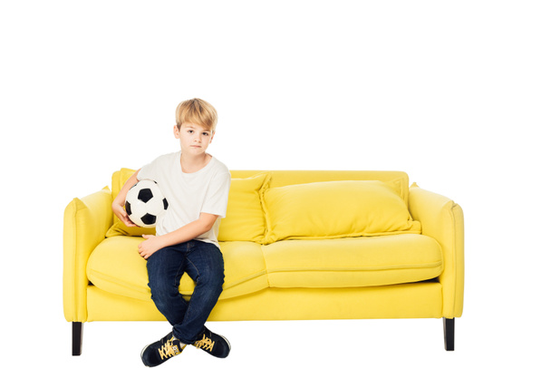 adorable boy sitting with football ball on yellow sofa isolated on white and looking at camera - Photo, Image