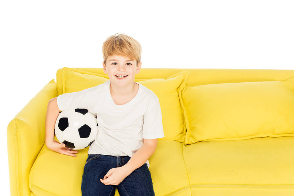smiling adorable boy sitting with football ball on yellow sofa isolated on white and looking at camera - Valokuva, kuva