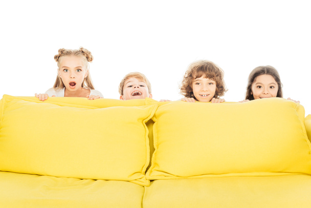 ενθουσιασμένοι και χαμογελαστά παιδιά κοιτάζοντας έξω από κίτρινο καναπέ και να βλέπουν τα φωτογραφικών μηχανών που απομονώνονται σε λευκό - Φωτογραφία, εικόνα