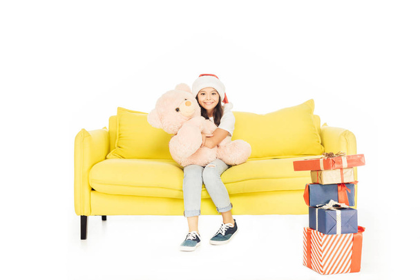 adorable child in santa hat hugging teddy bear on yellow sofa and looking at camera isolated on white - Photo, Image