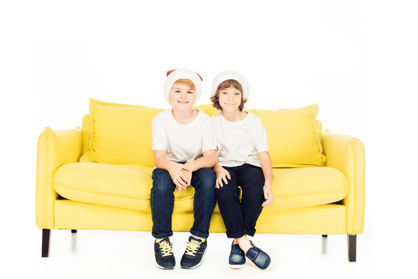 adorable boys in santa hats sitting on yellow sofa isolated on white, looking at camera - Foto, Imagen