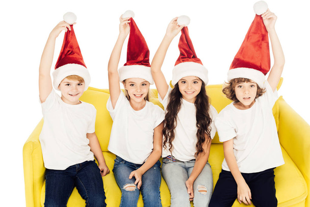 smiling adorable kids holding santa hats and sitting on yellow sofa isolated on white - Photo, image