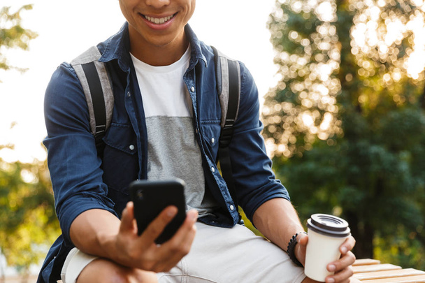 Cropped image of Smiling asian male student using smartphone and drinking coffee while sitting in park - Photo, Image