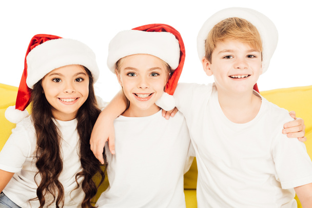 portrait of smiling adorable kids in santa hats sitting on yellow sofa and looking at camera isolated on white - Photo, Image
