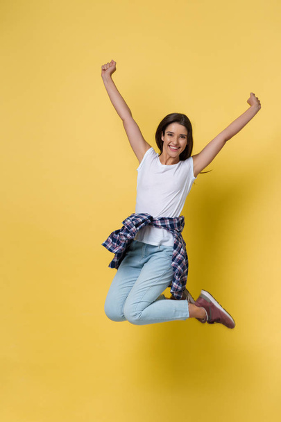 Full-length portrait of carefree girl in white shirt and jean jumping on yellow background. - Φωτογραφία, εικόνα
