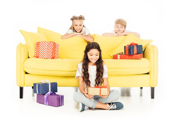 smiling adorable child holding gift box isolated on white, friends looking out from yellow sofa - Photo, image