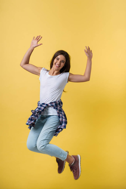 Full-length portrait of carefree girl in white shirt and jean jumping on yellow background. - Фото, изображение