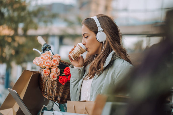 Mujer joven feliz teniendo una videollamada con un ordenador portátil y auriculares mientras está sentado en el banco y beber café
  - Foto, Imagen
