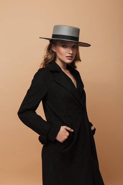 Jeune belle femme aux cheveux ondulés en manteau noir et chapeau regardant attentivement dans la caméra tenant la main dans des poches sur fond beige
 - Photo, image