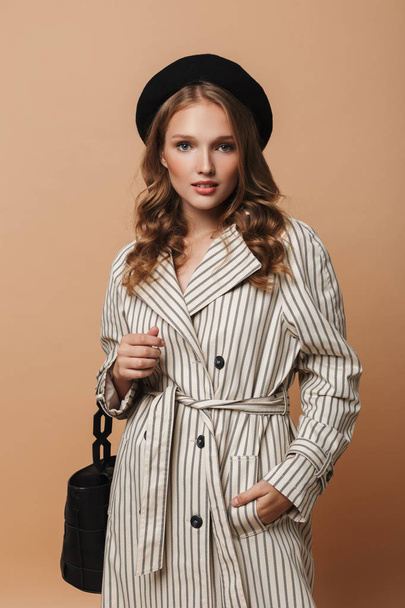 Young pretty woman with wavy hair in striped coat and black hat with handbag dreamily looking in camera over beige background - Photo, Image