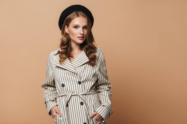 Young gorgeous woman in striped coat and black hat dreamily looking in camera holding hands in pockets over beige background - Zdjęcie, obraz