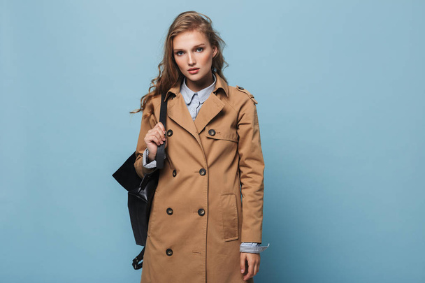 Beautiful girl with wavy hair in trench coat with backpack dreamily looking in camera over blue background - Фото, зображення
