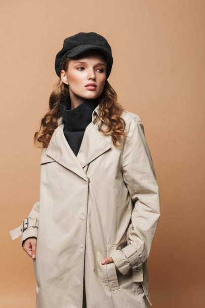 Young beautiful woman with wavy hair in trench coat and cap thoughtfully looking aside holding hands in pockets over beige background - Fotoğraf, Görsel