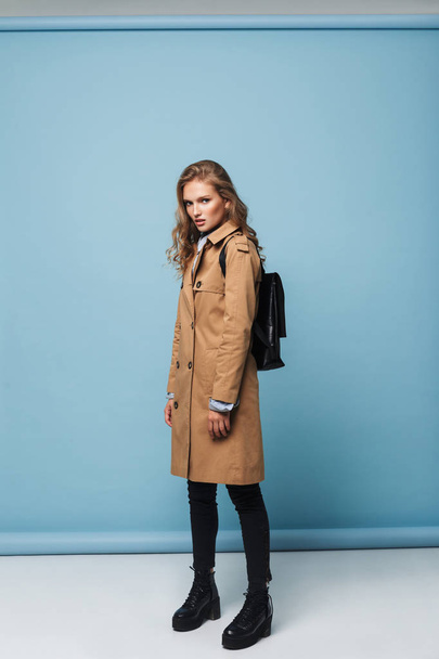 Young attractive woman with wavy hair in trench coat with black backpack thoughtfully looking in camera over blue background - Foto, afbeelding