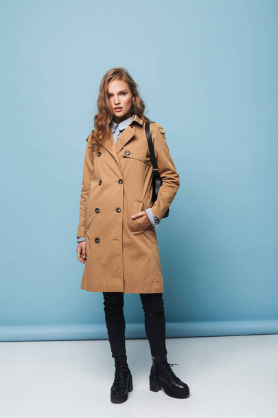 Young pensive woman with wavy hair in trench coat with backpack dreamily looking in camera over blue background - Foto, afbeelding