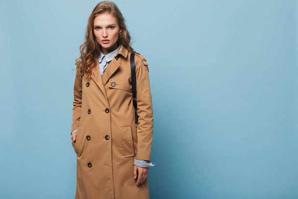 Young gorgeous woman with wavy hair in trench coat thoughtfully looking in camera over blue background - Photo, Image