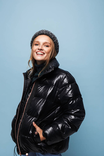 Young cheerful woman in knitted hat and black down jacket joyfully looking in camera holding hands in pockets over blue background - Photo, Image