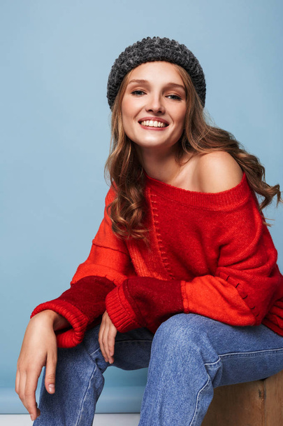 fröhliches Mädchen mit welligem Haar in Hut und rotem Pullover, das glücklich in die Kamera über blauem Hintergrund blickt - Foto, Bild