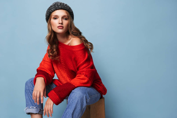 Young attractive woman with wavy hair in hat and red sweater thoughtfully looking in camera over blue background - Photo, Image