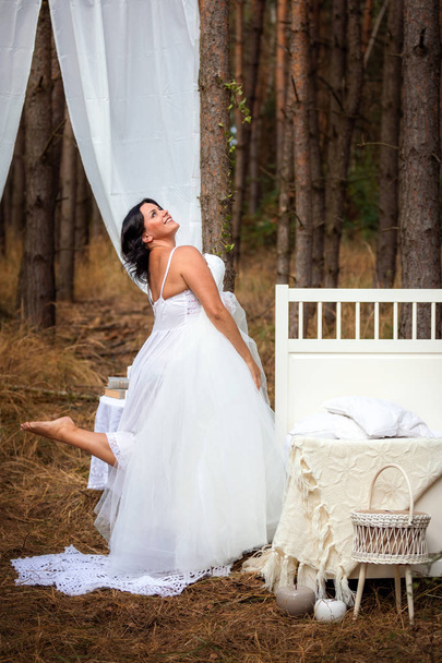 Vrouw probeert op trouwjurk naast bed in de ochtend op de achtergrond van de natuur van bos, ochtend van het concept van de bruid - Foto, afbeelding