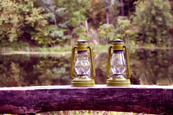 Lámpara de queroseno. El concepto de dos lámparas de aceite de pie de madera en el bosque contra el telón de fondo del lago. Sendero de emoción. Estilo rústico, boho
. - Foto, Imagen