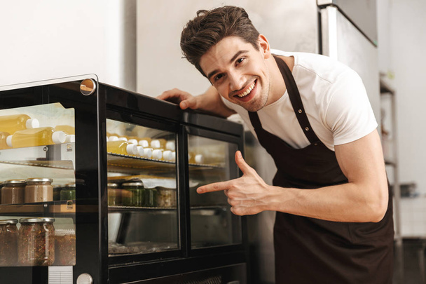 Image of happy excited young man chef on the kitchen pointing to spices and sauses. - Foto, immagini
