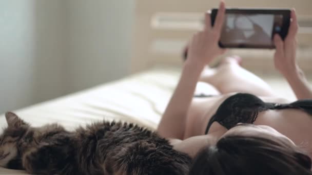 Young beautiful woman in black lingerie making selfie photo on a tablet lying on a bed nearby cat - Felvétel, videó