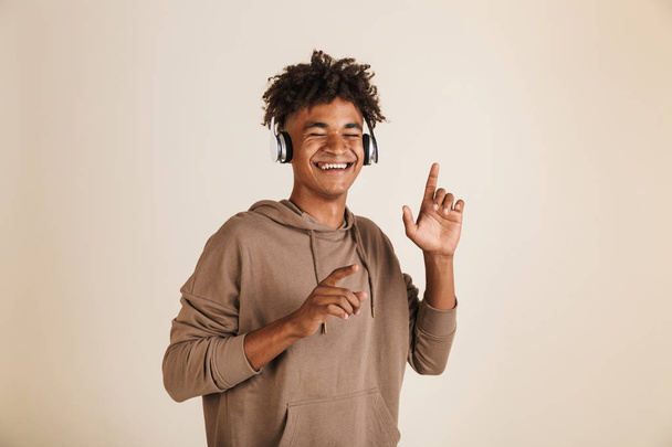 Portrait of a smiling young afro american man dressed in hoodie isolated, listening to music with headphones, dancing - Zdjęcie, obraz