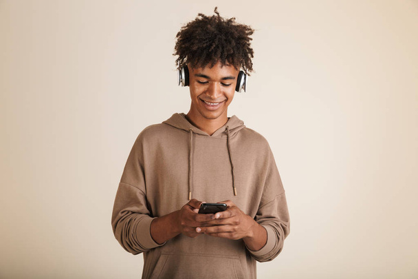 Portrait of a happy young afro american man dressed in hoodie isolated, listening to music with headphones, holding mobile phone - Foto, imagen