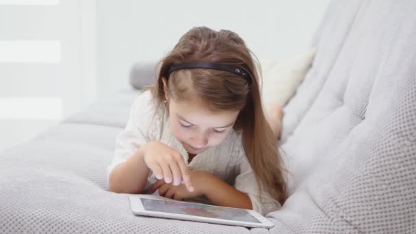 Pretty little girl lying on sofa using tablet . Small child playing on the laptop on an grey couch at home - Video, Çekim