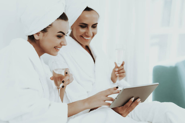 Girls In A Bathrobes Are Looking At Tablet Screen. Beautiful Shiny Smile. Sunny Day. Good Mood. Young And Attractive. Towel On Head. Good Mood. Drinking Champagne. Pre-wedding Care Concept. - Valokuva, kuva