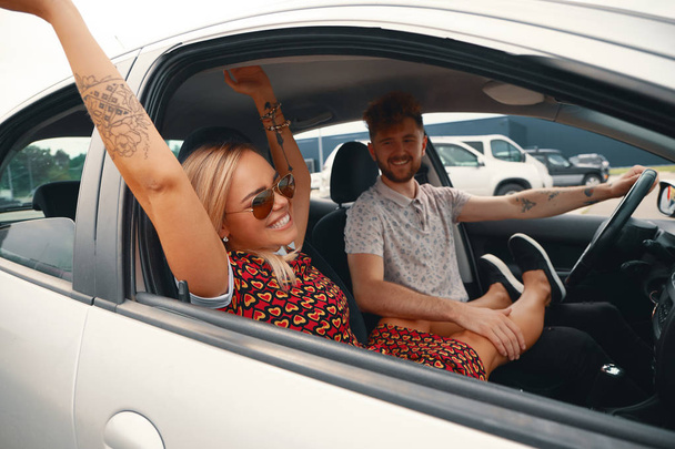 Onnellinen pari nauttimassa pitkästä ajomatkasta autossa. Ystävät ajavat automatkalla kesäpäivänä. Valkoihoinen nuori mies ajaa autoa ja iloinen nainen kädet ylhäällä
. - Valokuva, kuva