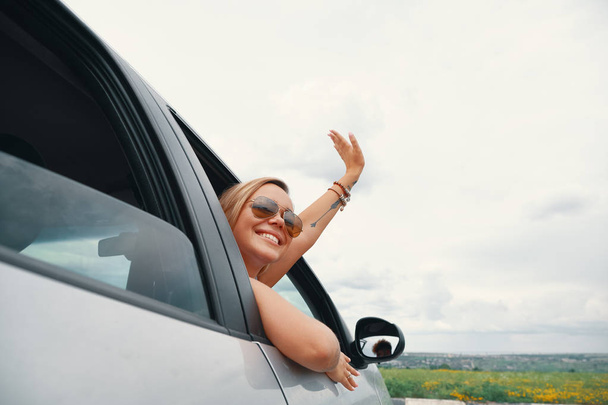 glückliche junge Frau genießt die Aussicht auf einer Roadtrip, während sie durch das Autofenster schaut und grüßt winkend ihre Hand. - Foto, Bild
