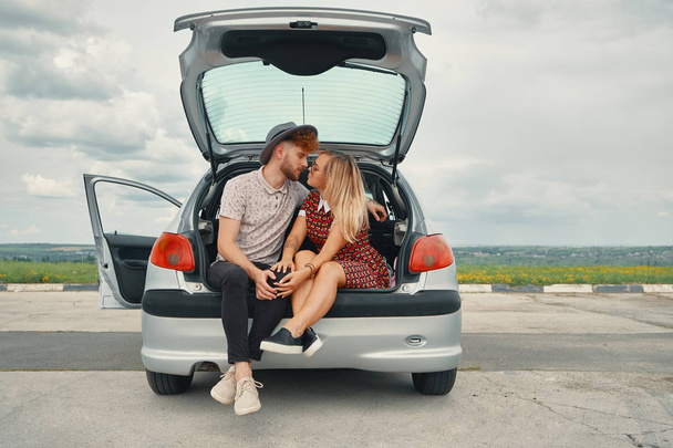 Beautiful carefree couple discovering the world traveling by car. Young man and woman kissing, hugging and touching each other while sitting in the car trunk - Photo, Image