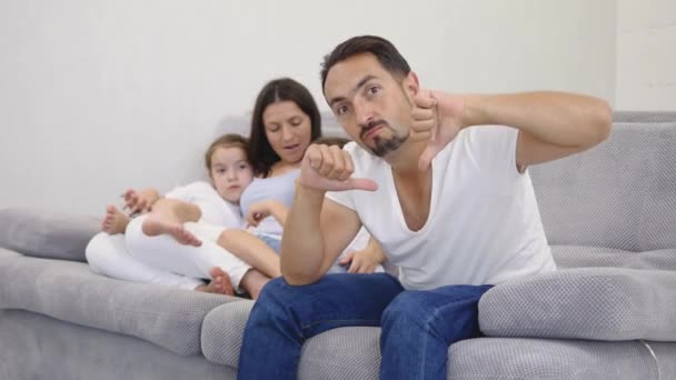 Сумний чоловік на передньому плані показує великі пальці вниз дружині і двом дітям в спині, сидячи на дивані
. - Кадри, відео