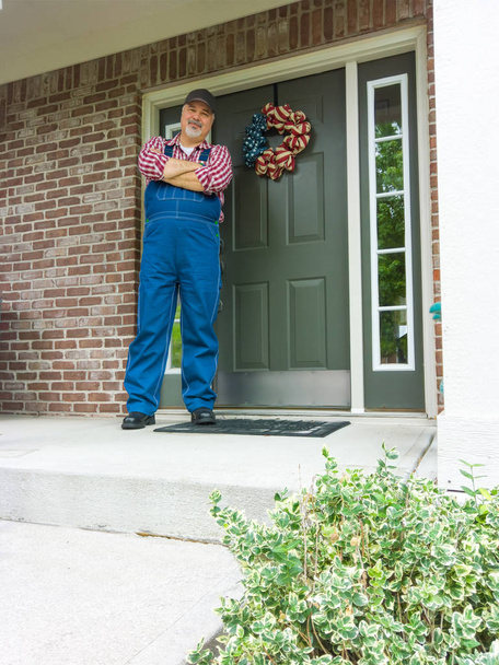 Feliz granjero con ropas patrióticas rojas, azules y blancas de pie en su porche el 4 de julio frente a una corona ornamental que celebra el Día de la Independencia.
 - Foto, imagen